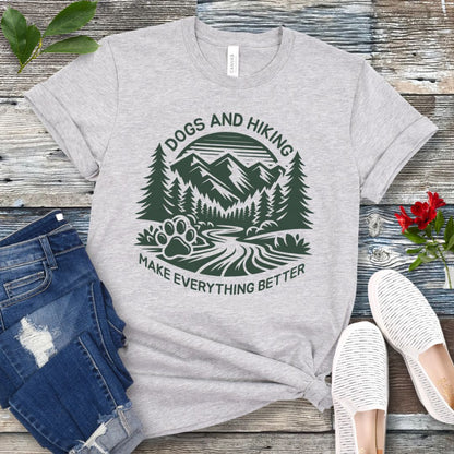 Gray t-shirt with mountains graphic with paw print and the text 'Dogs and Hiking Make Everything Better', shown on a wooden surface with jeans and shoes.