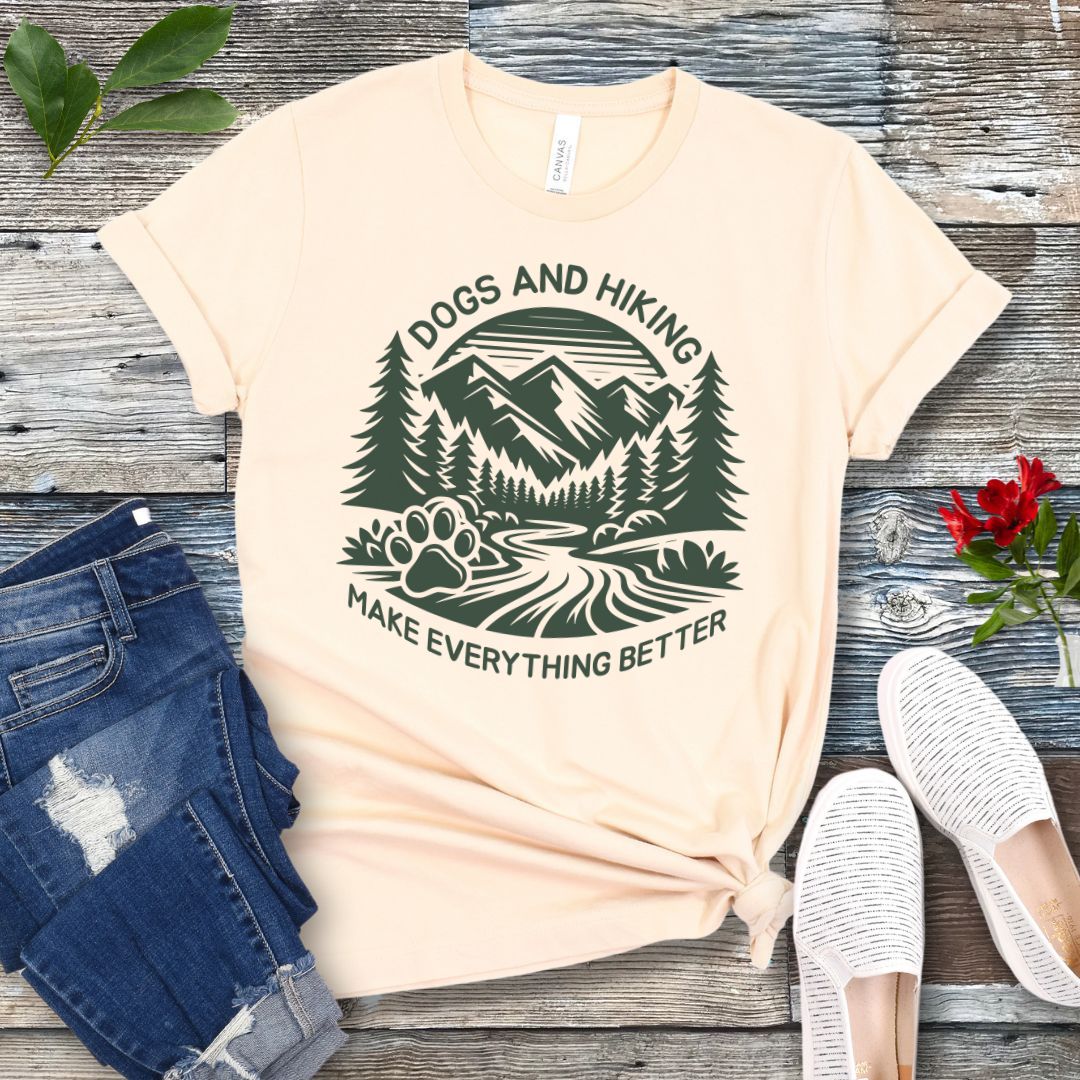 Beige t-shirt with mountains graphic with paw print and the text 'Dogs and Hiking Make Everything Better', shown on a wooden surface with jeans and shoes.