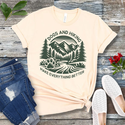 Beige t-shirt with mountains graphic with paw print and the text 'Dogs and Hiking Make Everything Better', shown on a wooden surface with jeans and shoes.