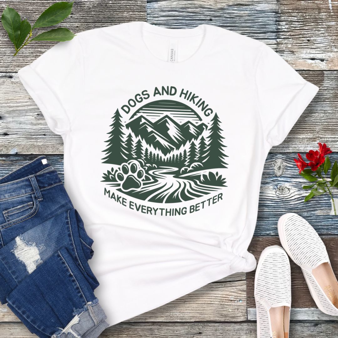 White t-shirt with mountains graphic with paw print and the text 'Dogs and Hiking Make Everything Better', shown on a wooden surface with jeans and shoes.