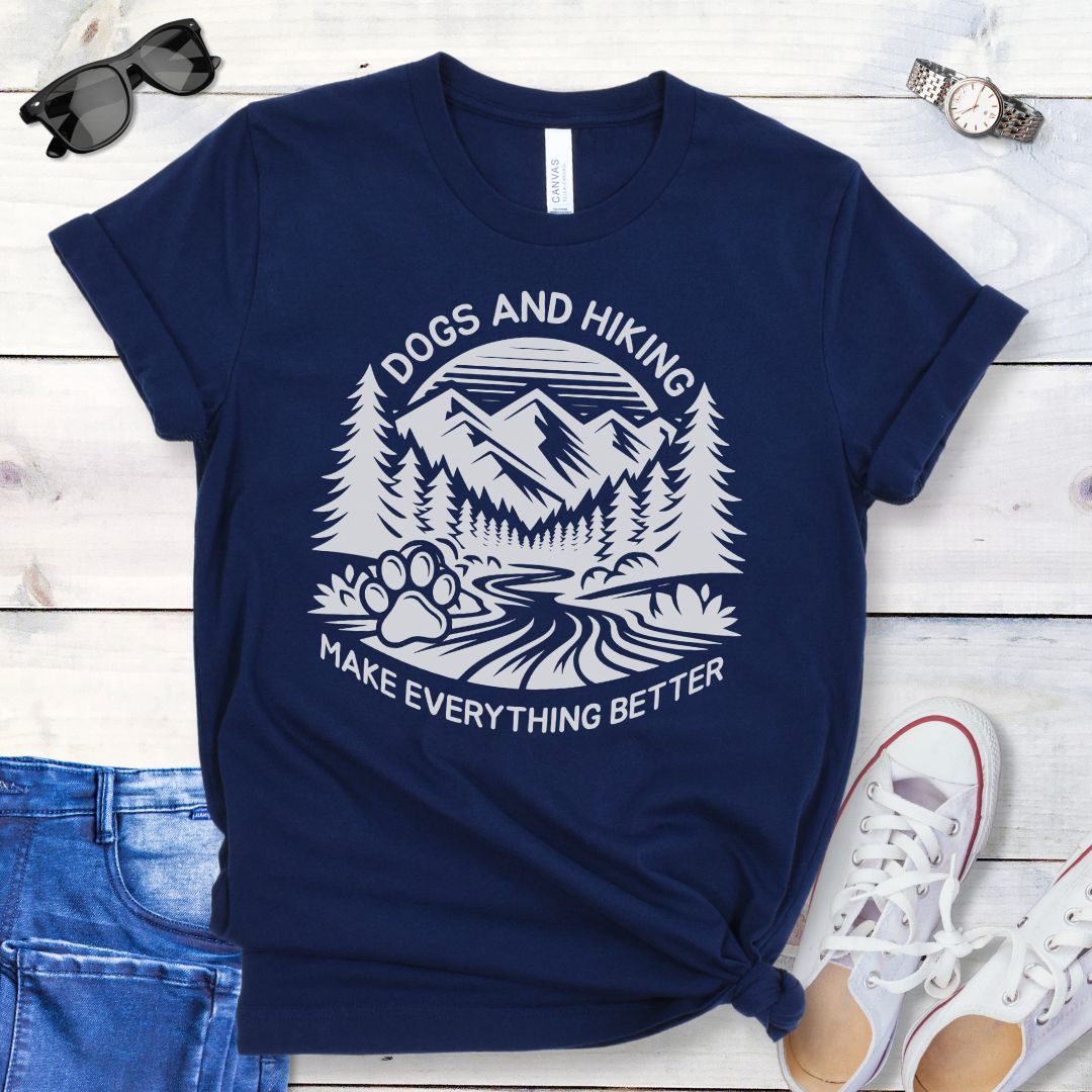 Navy blue t-shirt with mountains graphic with paw print and the text 'Dogs and Hiking Make Everything Better', shown on a wooden surface with jeans and shoes.