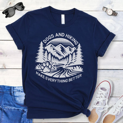 Navy blue t-shirt with mountains graphic with paw print and the text 'Dogs and Hiking Make Everything Better', shown on a wooden surface with jeans and shoes.