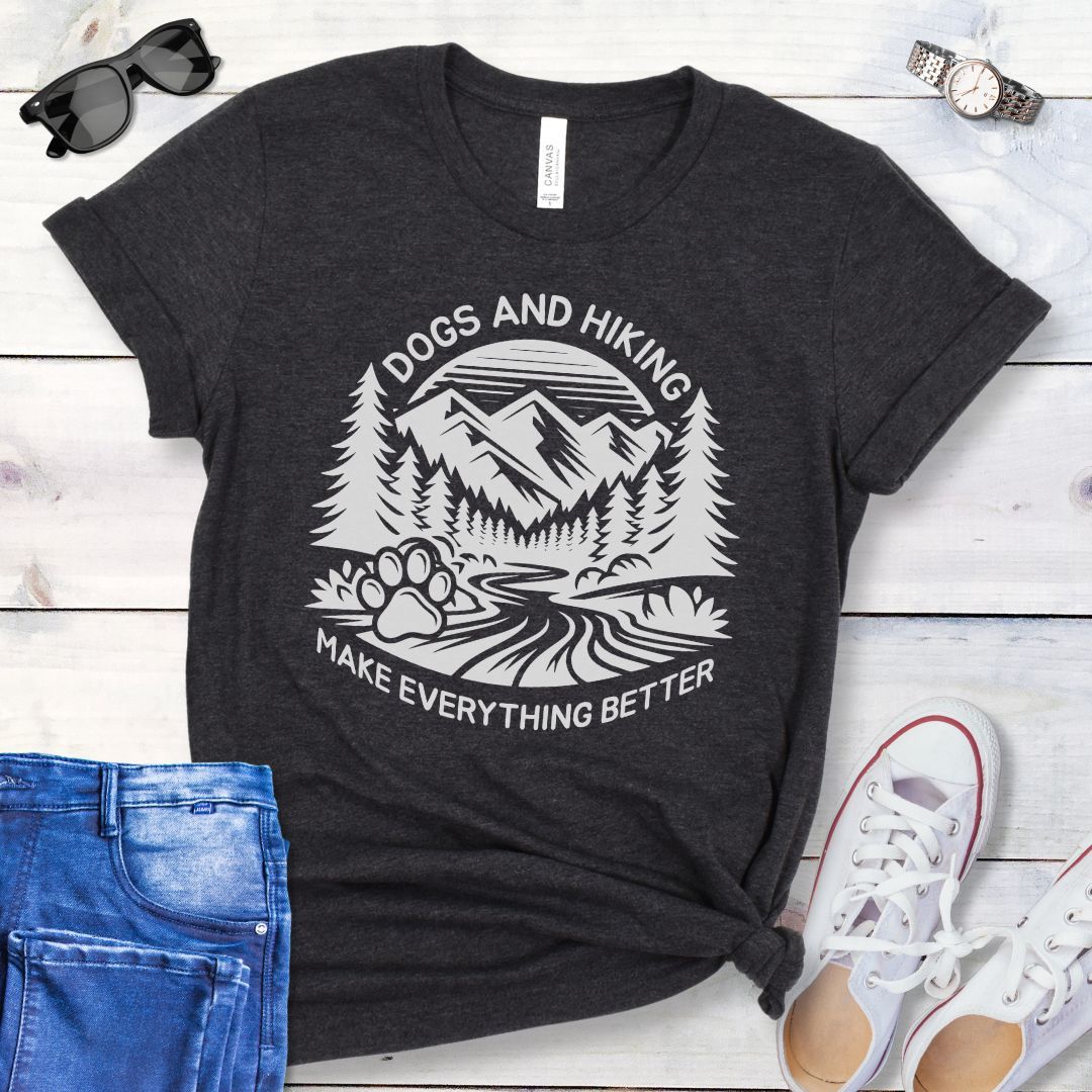 Dark gray t-shirt with mountains graphic with paw print and the text 'Dogs and Hiking Make Everything Better', shown on a wooden surface with jeans and shoes.