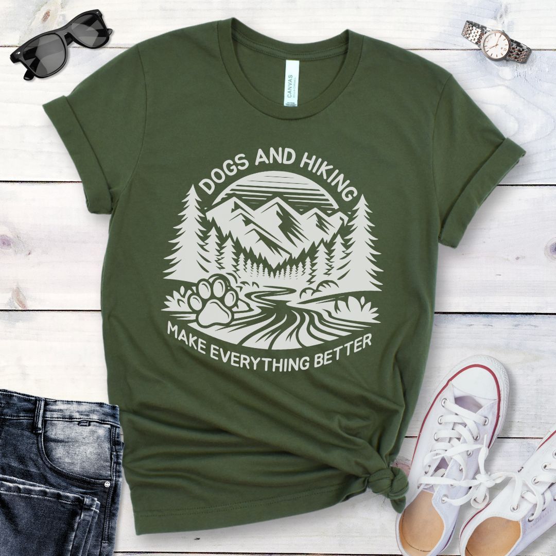 Military green t-shirt with mountains graphic with paw print and the text 'Dogs and Hiking Make Everything Better', shown on a wooden surface with jeans and shoes.