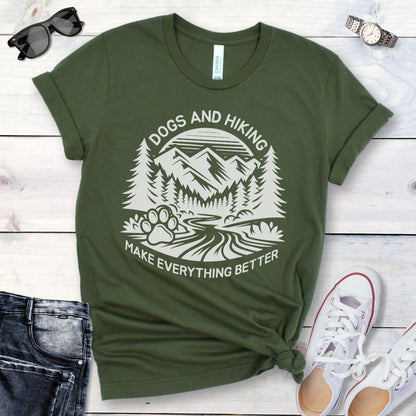 Military green t-shirt with mountains graphic with paw print and the text 'Dogs and Hiking Make Everything Better', shown on a wooden surface with jeans and shoes.
