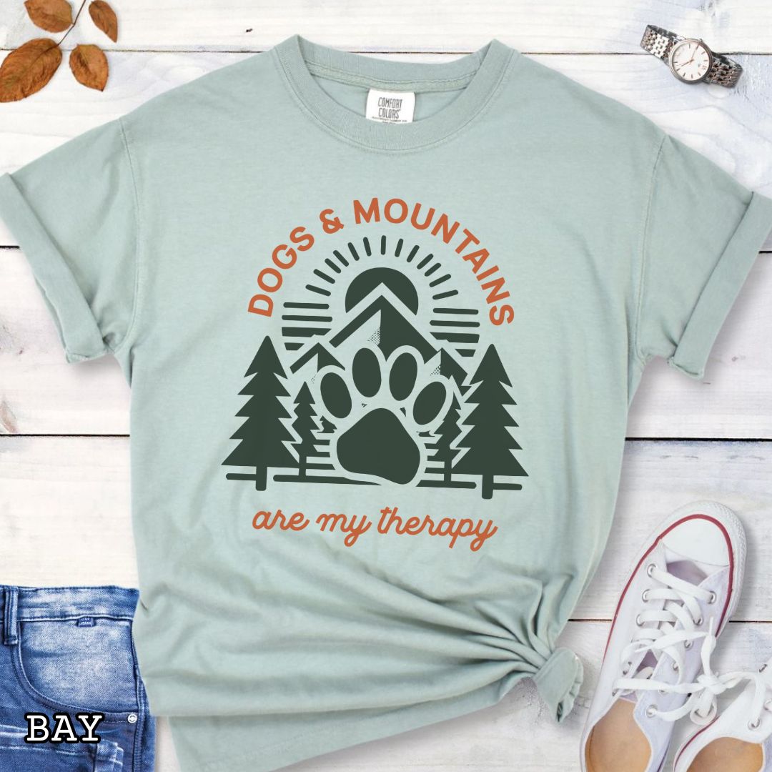 Light green Comfort Colors t-shirt for outdoorsy dog lovers with graphic of mountains, trees and dog paw, and the text 'Dogs & Mountains are my therapy', shown on a wooden surface with jeans and flowers.