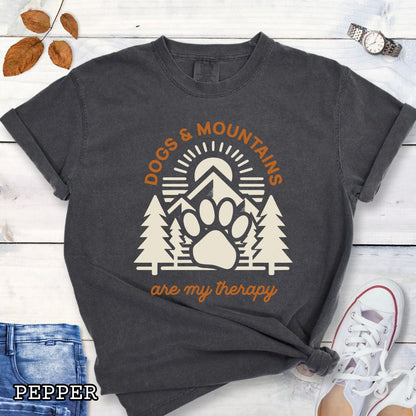 Dark grey Comfort Colors t-shirt for outdoorsy dog lovers with graphic of mountains, trees and dog paw, and the text 'Dogs & Mountains are my therapy', shown on a wooden surface with jeans and flowers.