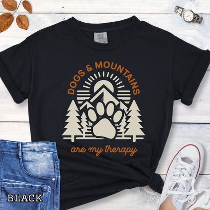 Black Comfort Colors t-shirt for outdoorsy dog lovers with graphic of mountains, trees and dog paw, and the text 'Dogs & Mountains are my therapy', shown on a wooden surface with jeans and flowers.