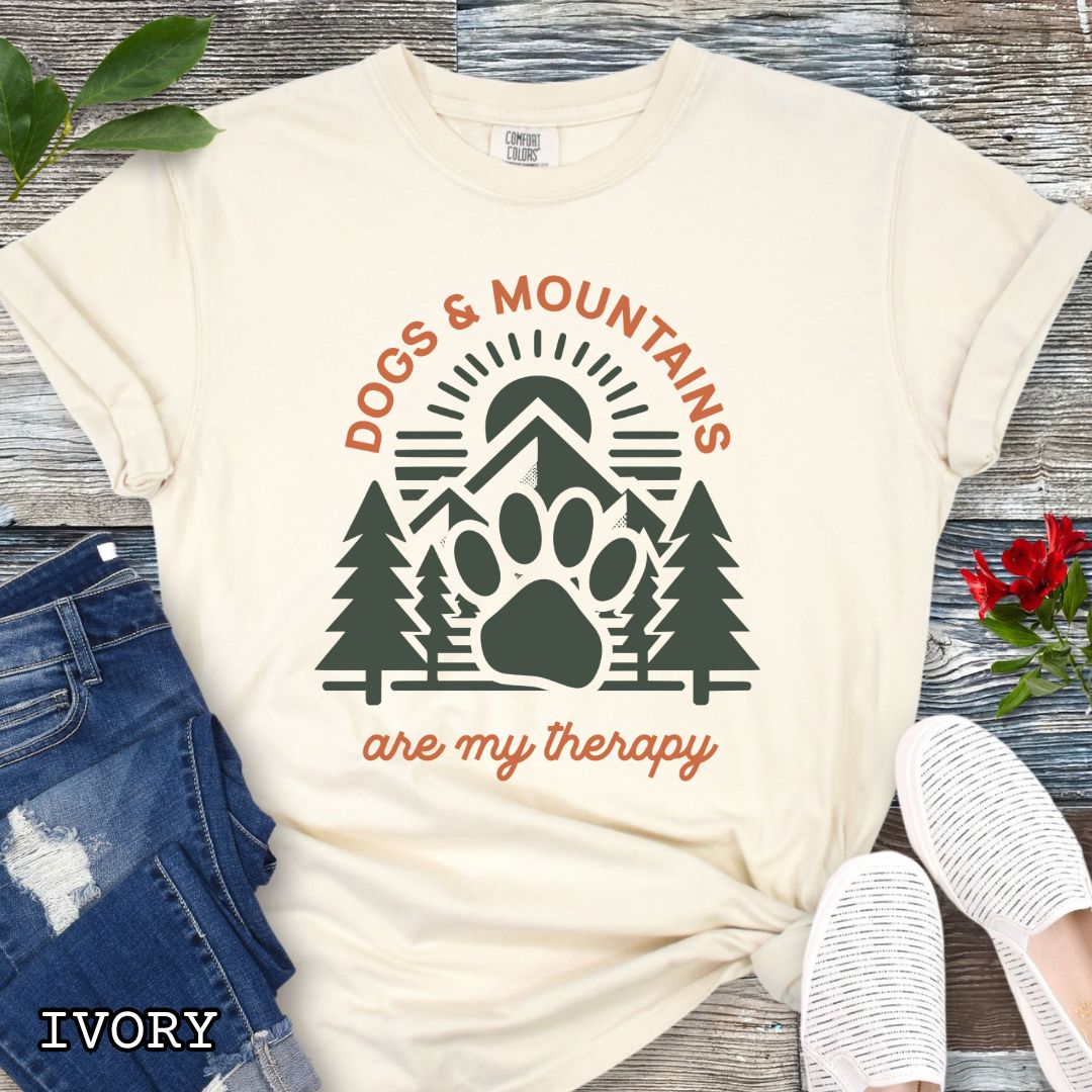 Beige Comfort Colors t-shirt for outdoorsy dog lovers with graphic of mountains, trees and dog paw, and the text 'Dogs & Mountains are my therapy', shown on a wooden surface with jeans and flowers.