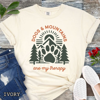 Beige Comfort Colors t-shirt for outdoorsy dog lovers with graphic of mountains, trees and dog paw, and the text 'Dogs & Mountains are my therapy', shown on a wooden surface with jeans and flowers.
