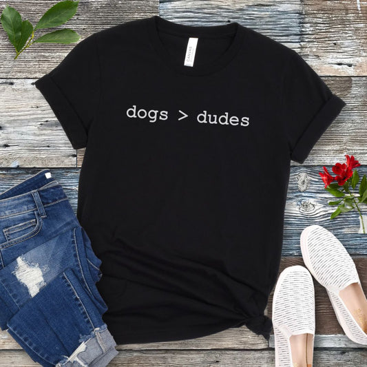 Black t-shirt for dog lovers with humorous text 'dogs > dudes' in typewriter style lettering, shown on a wooden surface with jeans and shoes.