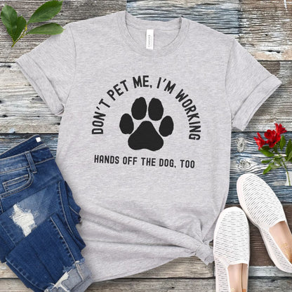 A grey T-shirt with the text 'DON'T PET ME, I'M WORKING. HANDS OFF THE DOG, TOO' and a paw print design, displayed on a wooden surface with a pair of jeans and white shoes in the background.