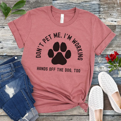 A mauve T-shirt with the text 'DON'T PET ME, I'M WORKING, HANDS OFF THE DOG, TOO' and a paw print design, displayed on a wooden surface with a pair of jeans and white shoes in the background.