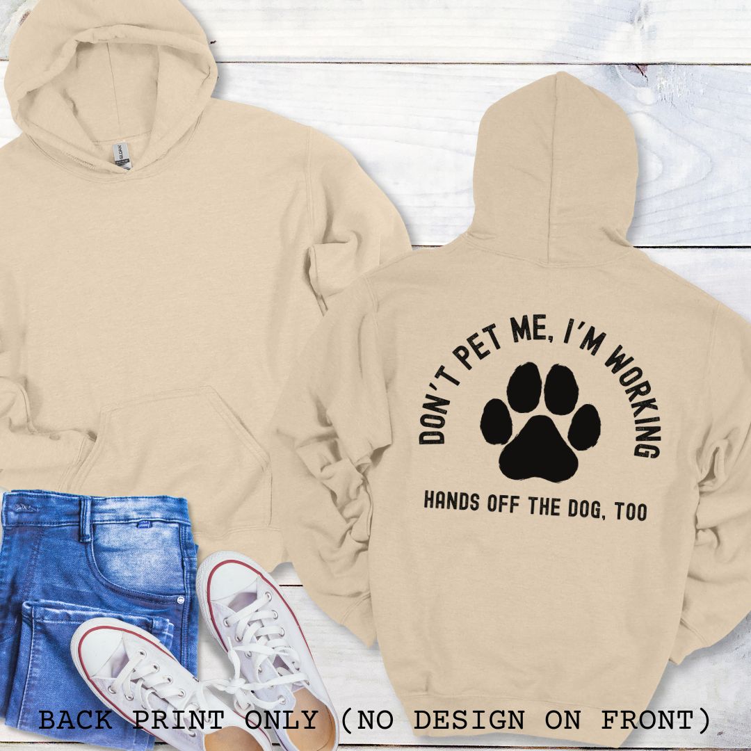 A sand colored back print hoodie with the text 'DON'T PET ME, I'M WORKING, HANDS OFF THE DOG, TOO' and a paw print design, displayed on a wooden surface with a pair of jeans and white shoes in the background.