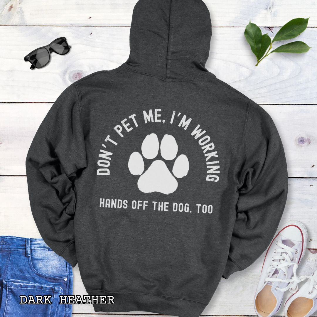 Dark grey back print hoodie with the text 'DON'T PET ME, I'M WORKING, HANDS OFF THE DOG, TOO' and a paw print design, displayed on a wooden surface with a pair of jeans and white shoes in the background.