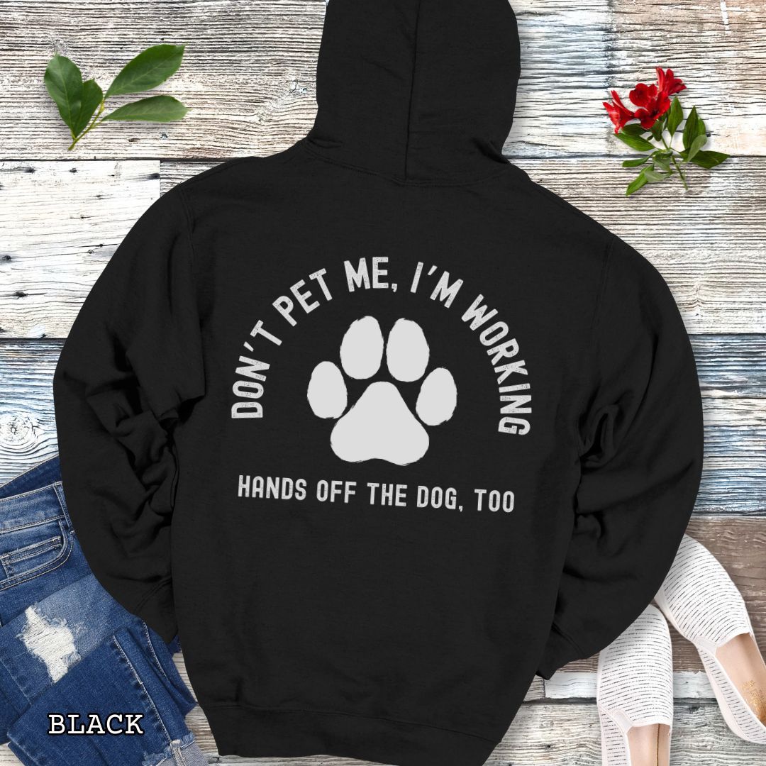 Black back print hoodie with the text 'DON'T PET ME, I'M WORKING, HANDS OFF THE DOG, TOO' and a paw print design, displayed on a wooden surface with a pair of jeans and white shoes in the background.