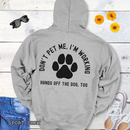 Medium grey back print hoodie with the text 'DON'T PET ME, I'M WORKING, HANDS OFF THE DOG, TOO' and a paw print design, displayed on a wooden surface with a pair of jeans and white shoes in the background.