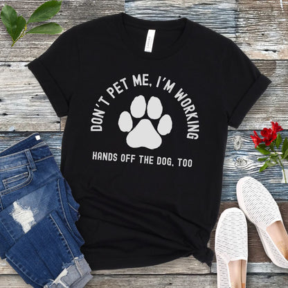 A black T-shirt with the text 'DON'T PET ME, I'M WORKING. HANDS OFF THE DOG, TOO' and a paw print design, displayed on a wooden surface with a pair of jeans and white shoes in the background.