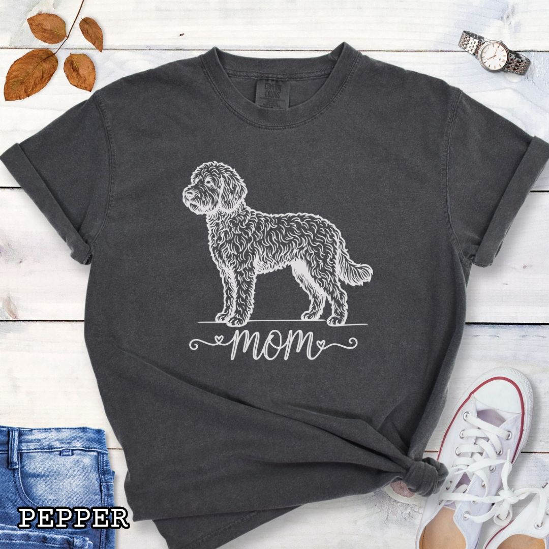 Dark grey Comfort Colors t-shirt for Doodle dog moms with a vintage-style Doodle dog illustration and 'mom' text in script lettering beneath it, shown on a wooden surface with jeans and shoes.