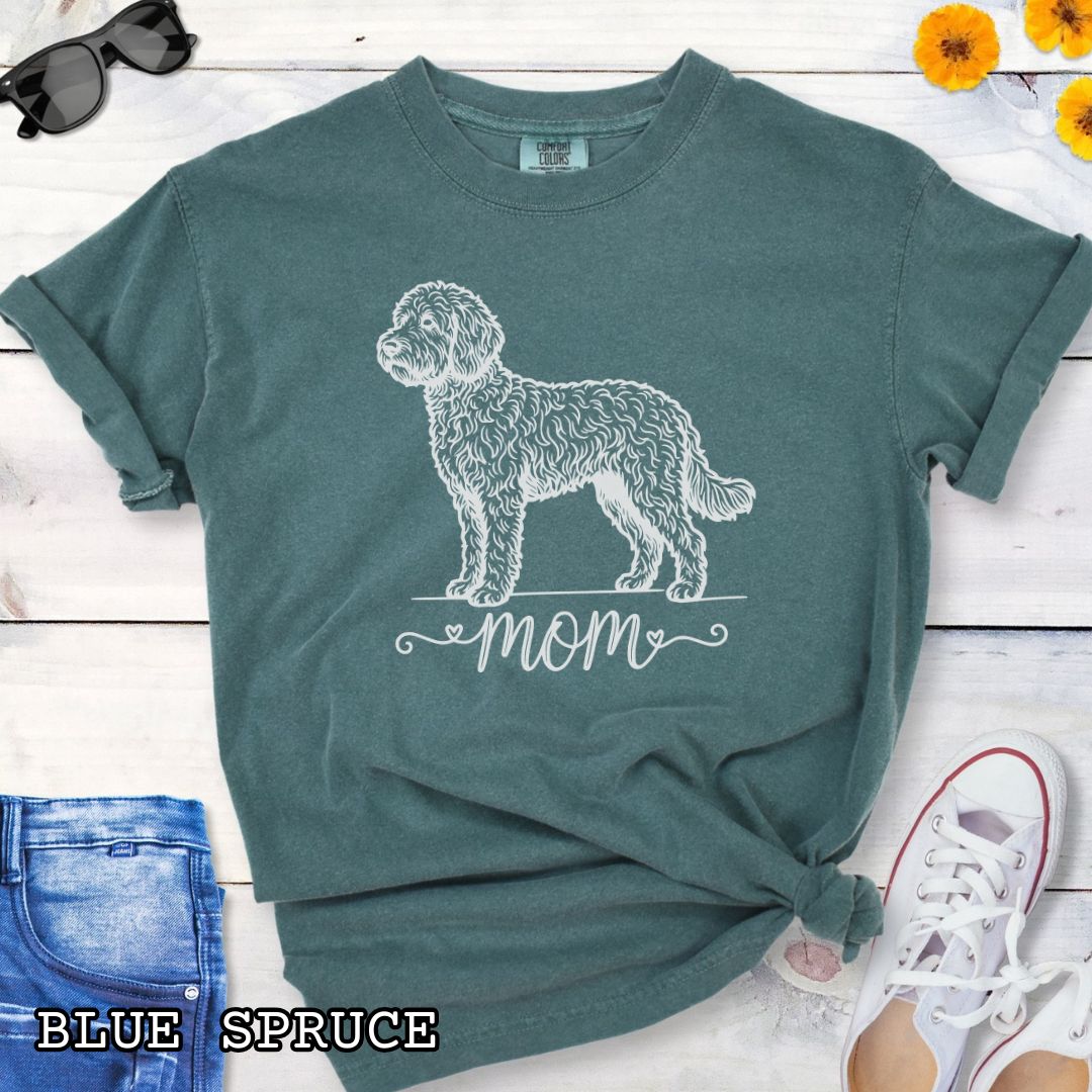 Green Comfort Colors t-shirt for Doodle dog moms with a vintage-style Doodle dog illustration and 'mom' text in script lettering beneath it, shown on a wooden surface with jeans and shoes.