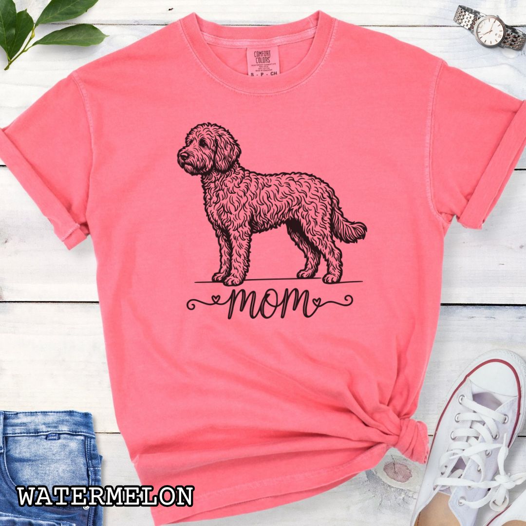 Pink Comfort Colors t-shirt for Doodle dog moms with a vintage-style Doodle dog illustration and 'mom' text in script lettering beneath it, shown on a wooden surface with jeans and shoes.