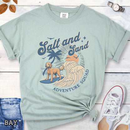 Light green Comfort Colors t-shirt for Labradoodle and Goldendoodle dog lovers with beach-themed graphic of a surfing Doodle dog, waves and sunset, and the text 'Salt and Sand Adventure Squad' in retro lettering, shown on a wooden surface with accessories.