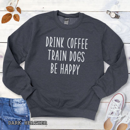 Dark grey unisex dog training sweatshirt with 'Drink Coffee Train Dogs Be Happy' text on a wooden surface with jeans and shoes.