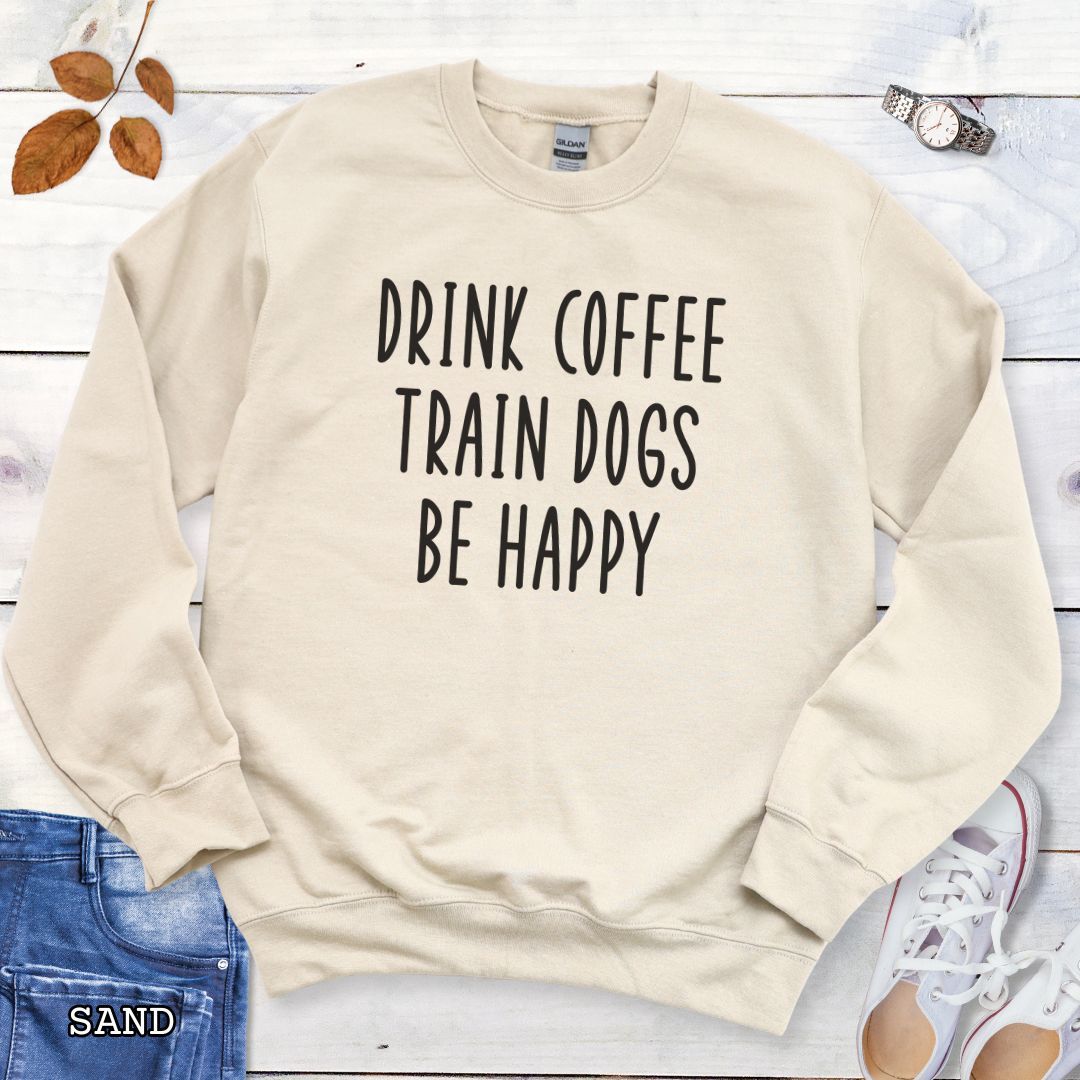 Beige unisex dog training sweatshirt with 'Drink Coffee Train Dogs Be Happy' text on a wooden surface with jeans and shoes.