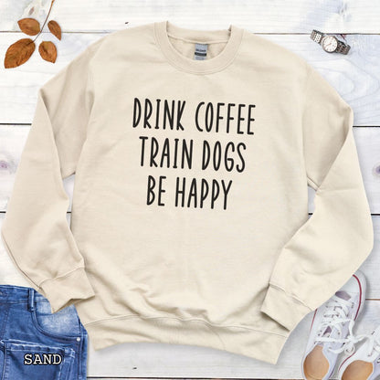 Beige unisex dog training sweatshirt with 'Drink Coffee Train Dogs Be Happy' text on a wooden surface with jeans and shoes.