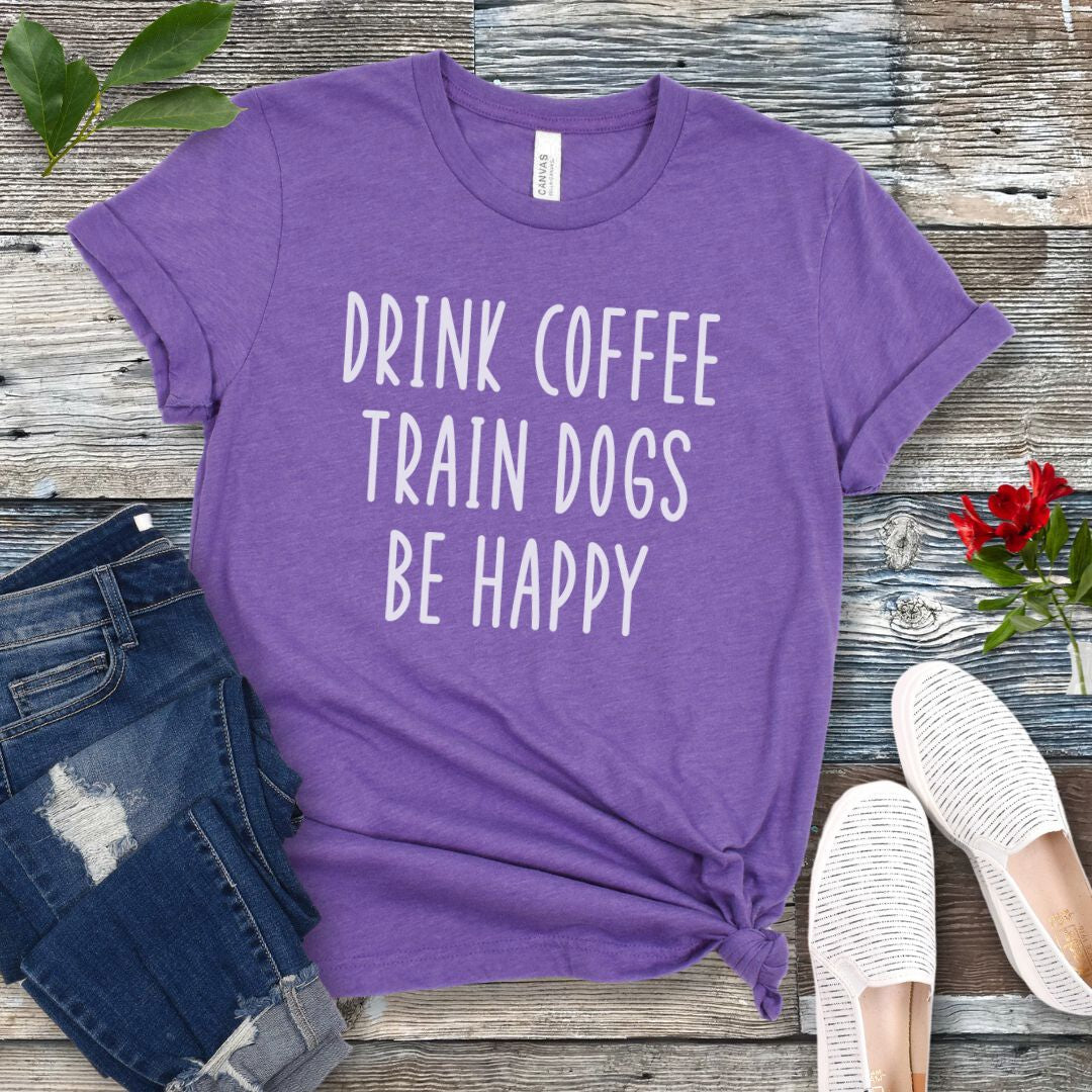 Purple unisex dog training t-shirt with 'Drink Coffee Train Dogs Be Happy' text on a wooden surface with jeans and shoes.