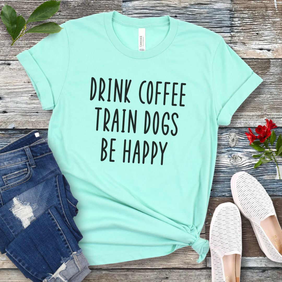 Mint green unisex dog training t-shirt with 'Drink Coffee Train Dogs Be Happy' text on a wooden surface with jeans and shoes.