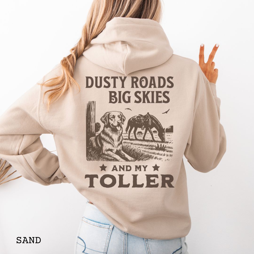 Back view of a woman wearing a beige hoodie featuring a Western-style design with grazing horse, rustic fence, and a Duck Tolling Retriever illustration; text reads “Dusty Roads, Big Skies, and My Toller.”