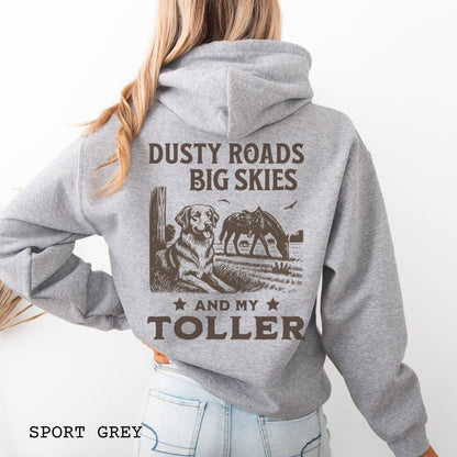 Back view of a woman wearing a grey hoodie featuring a Western-style design with grazing horse, rustic fence, and a Duck Tolling Retriever illustration; text reads “Dusty Roads, Big Skies, and My Toller.”