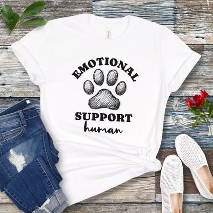 White unisex t-shirt for dog lovers with large paw print and humorous text 'EMOTIONAL SUPPORT human' in bold lettering, shown on a wooden surface alongside jeans and shoes.