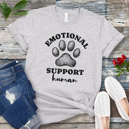 Grey unisex t-shirt for dog lovers with large paw print and humorous text 'EMOTIONAL SUPPORT human' in bold lettering, shown on a wooden surface alongside jeans and shoes.