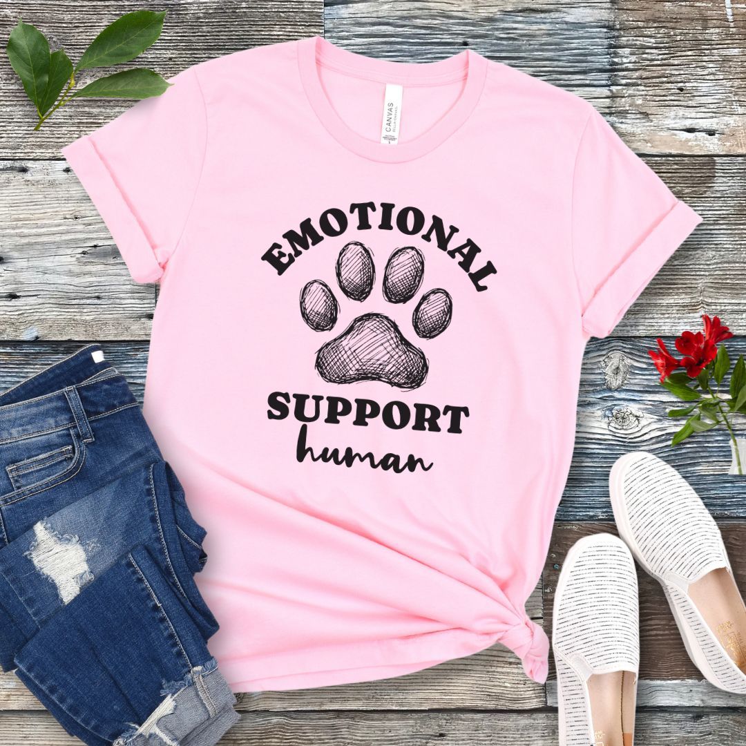 Pink unisex t-shirt for dog lovers with large paw print and humorous text 'EMOTIONAL SUPPORT human' in bold lettering, shown on a wooden surface alongside jeans and shoes.