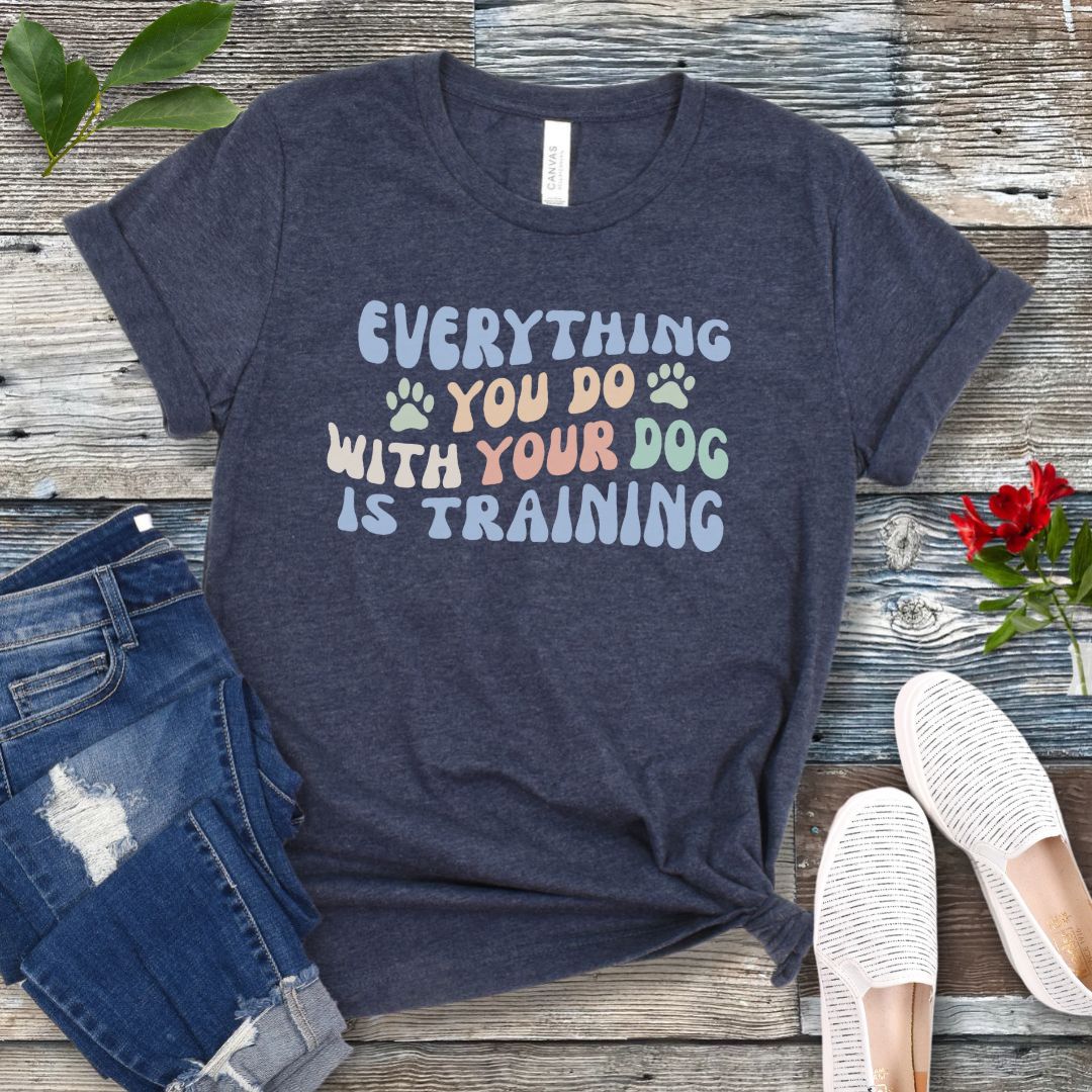 Heather navy blue dog training t-shirt with colorful text that reads "Everything you do with your dog is training" on a wooden surface with jeans and shoes.