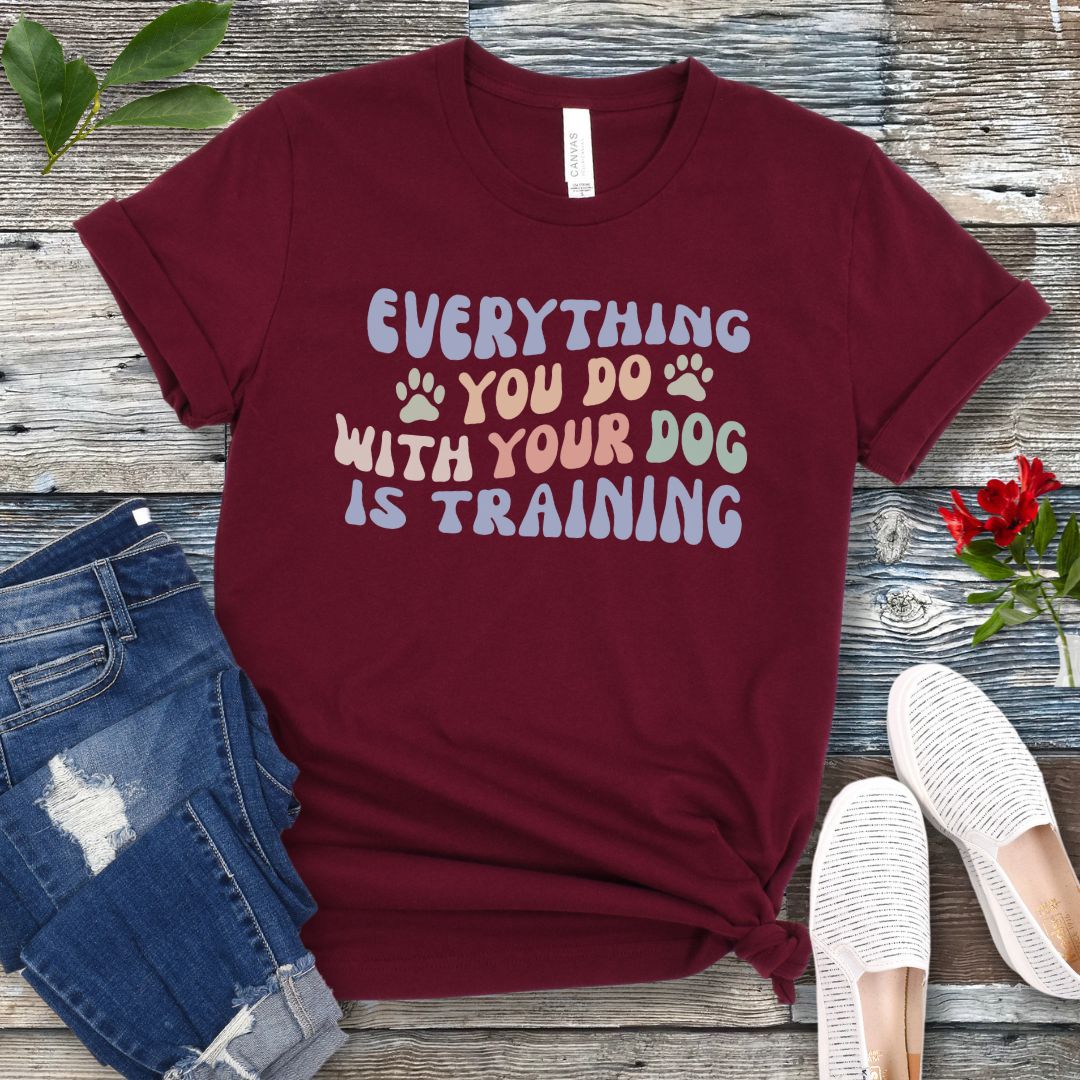 Maroon dog training t-shirt with colorful text that reads "Everything you do with your dog is training" on a wooden surface with jeans and shoes.