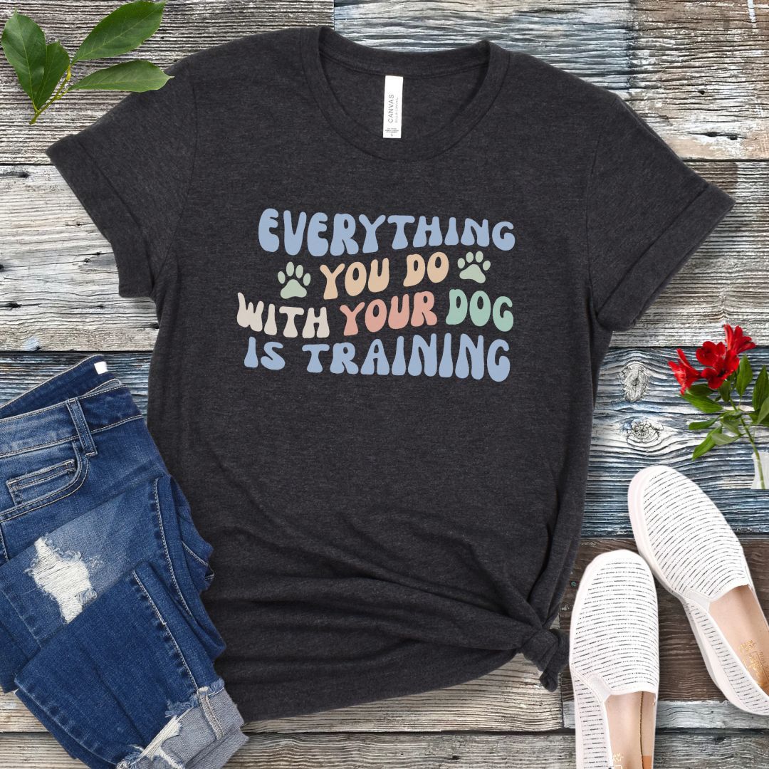 Dark grey dog training t-shirt with colorful text that reads "Everything you do with your dog is training" on a wooden surface with jeans and shoes.