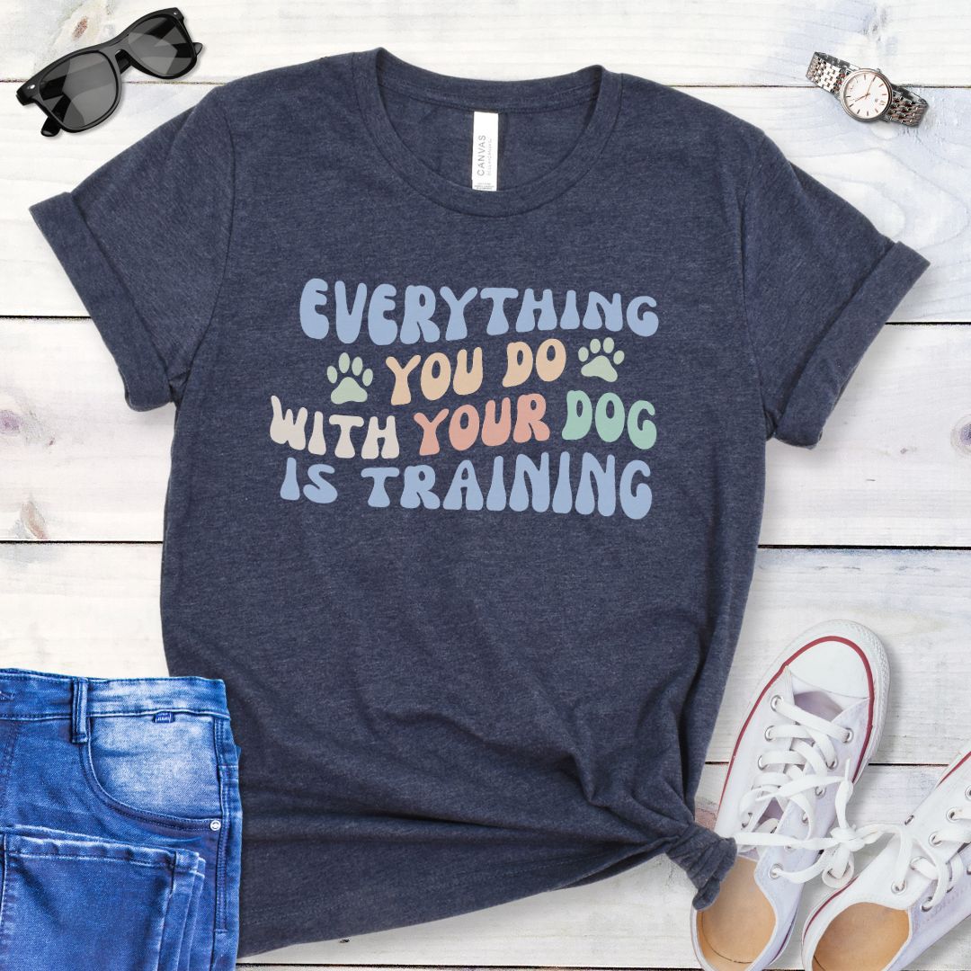 Heather navy dog training t-shirt with colorful retro style text that reads "Everything you do with your dog is training", shown on a wooden surface with jeans and shoes.