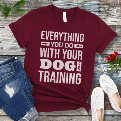 A maroon t-shirt with the text 'EVERYTHING YOU DO WITH YOUR DOG IS TRAINING' text design, displayed flat with a pair of blue jeans and white shoes.