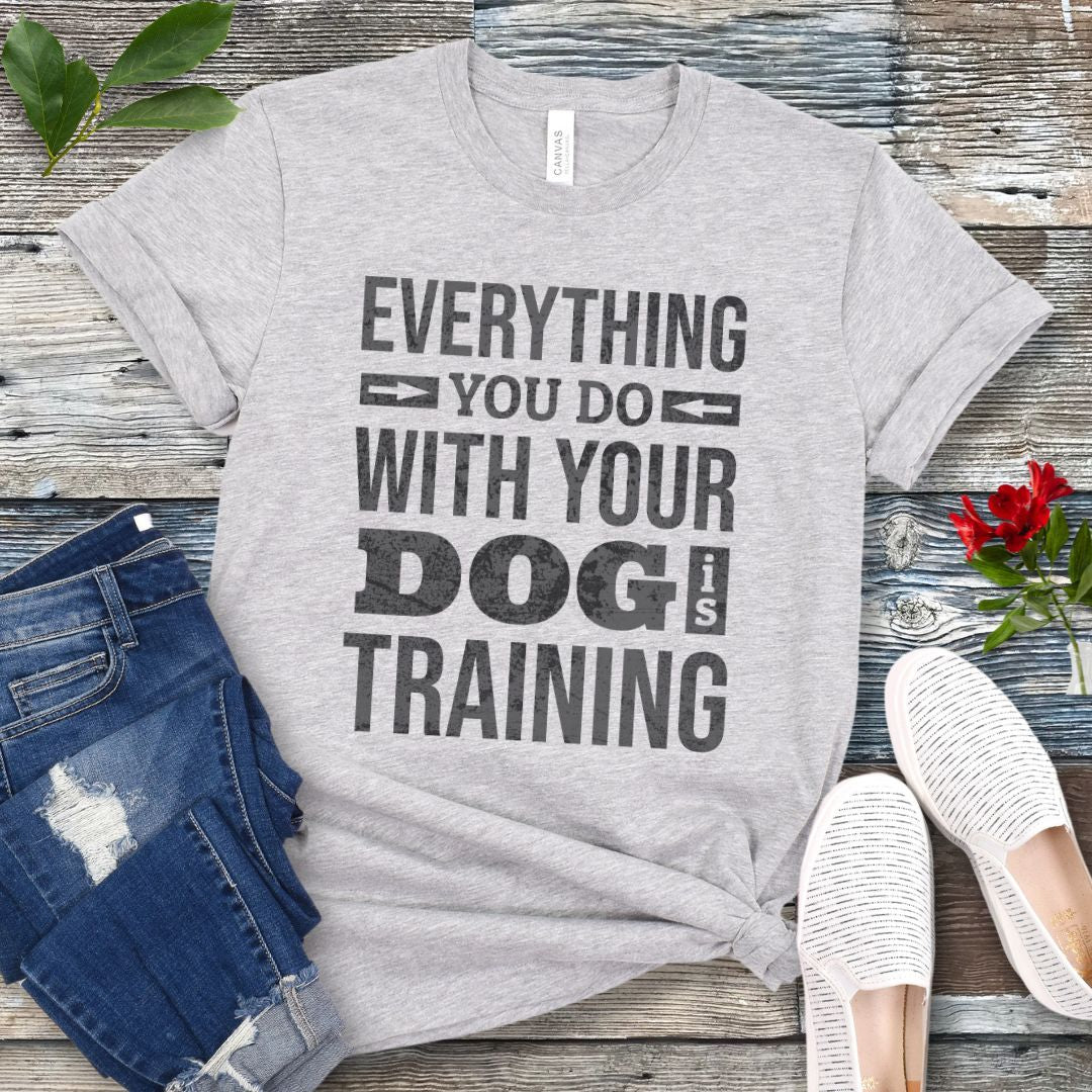 A heather gray graphic t-shirt with the text 'EVERYTHING YOU DO WITH YOUR DOG IS TRAINING' in distressed black lettering. The t-shirt is styled in a flat-lay arrangement on a weathered wood background, accompanied by blue jeans, white slip-on shoes, and a small red rose.