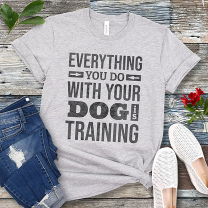 A heather gray graphic t-shirt with the text 'EVERYTHING YOU DO WITH YOUR DOG IS TRAINING' in distressed black lettering. The t-shirt is styled in a flat-lay arrangement on a weathered wood background, accompanied by blue jeans, white slip-on shoes, and a small red rose.