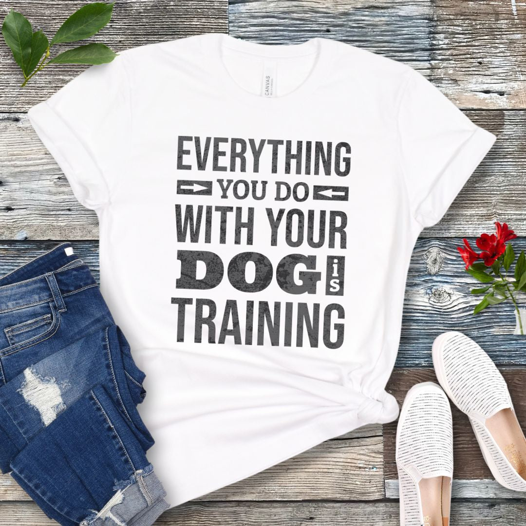 A white graphic t-shirt with the text 'EVERYTHING YOU DO WITH YOUR DOG IS TRAINING' printed in black distressed lettering. The shirt is styled in a flat-lay arrangement on a weathered gray wooden background, accompanied by blue denim shorts, white slip-on shoes, and a small red rose.