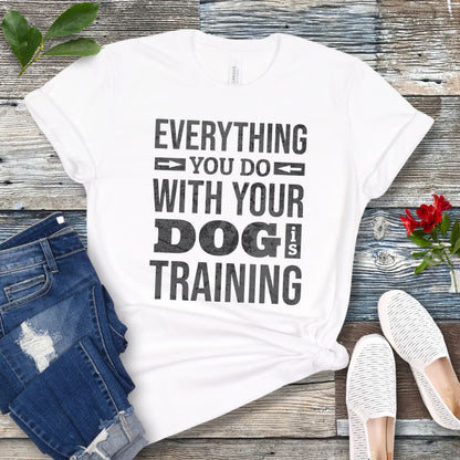 A white graphic t-shirt with the text 'EVERYTHING YOU DO WITH YOUR DOG IS TRAINING' printed in black distressed lettering. The shirt is styled in a flat-lay arrangement on a weathered gray wooden background, accompanied by blue denim shorts, white slip-on shoes, and a small red rose.
