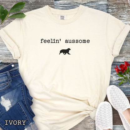 Beige Comfort Colors t-shirt for Australian Shepherd dog lovers with 'feelin' aussome' text in typewriter font and a trotting dog silhouette, shown on a white wooden surface with shoes and jeans.
