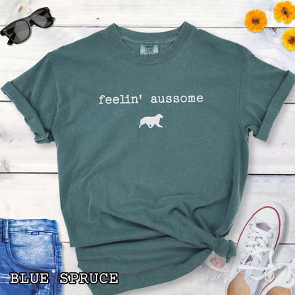 Green Comfort Colors t-shirt for Australian Shepherd dog lovers with 'feelin' aussome' text in typewriter font and a trotting dog silhouette, shown on a white wooden surface with shoes and jeans.
