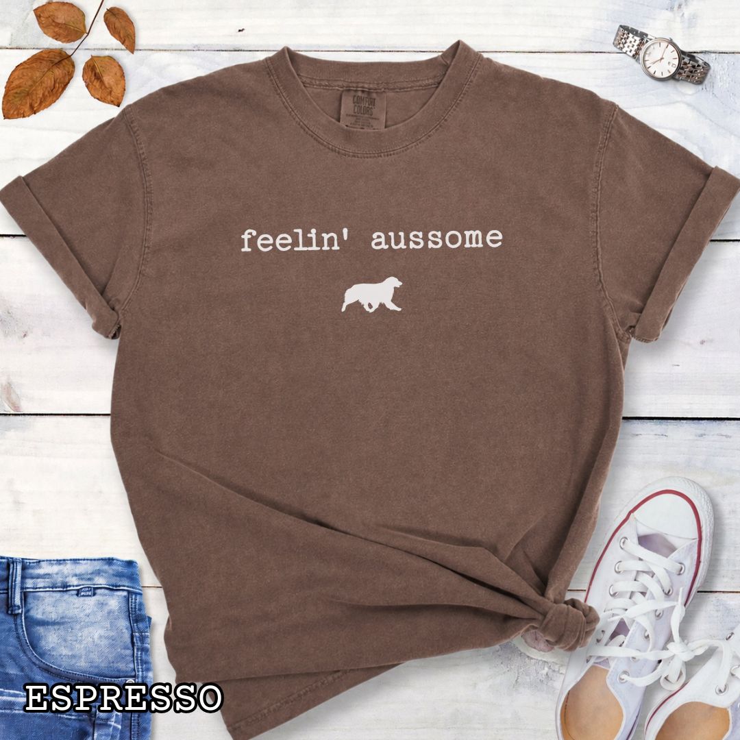 Brown Comfort Colors t-shirt for Australian Shepherd dog lovers with 'feelin' aussome' text in typewriter font and a trotting dog silhouette, shown on a white wooden surface with shoes and jeans.