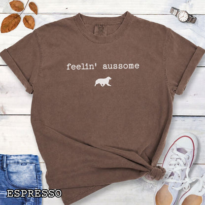 Brown Comfort Colors t-shirt for Australian Shepherd dog lovers with 'feelin' aussome' text in typewriter font and a trotting dog silhouette, shown on a white wooden surface with shoes and jeans.