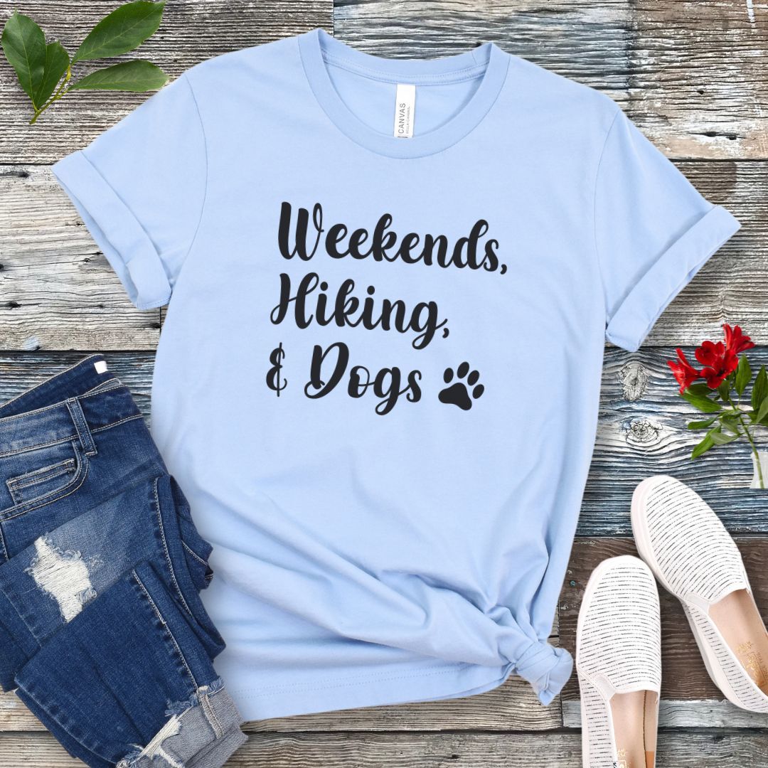 A light blue t-shirt with the text 'Weekends, Hiking, & Dogs' text in playful, feminine script and a paw print, displayed on a wooden surface alongside denim jeans and white sneakers.
